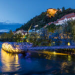 Blick von der Murbrücke auf die blau leuchtende Murinsel in Graz